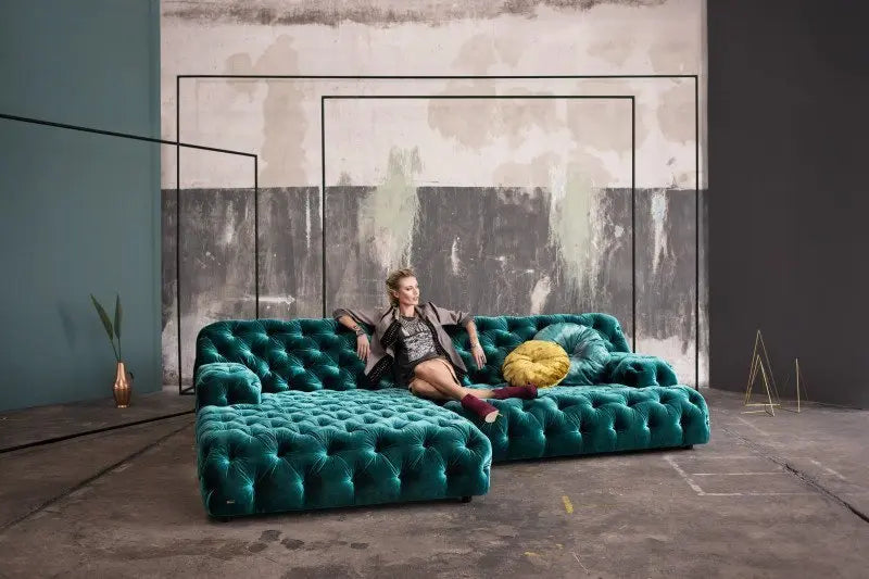 A person sits on the spacious, teal Coco Miralda Sofa, characterized by its tufted design and large sectional layout, positioned in a corner of an expansive, industrial-style room with unfinished walls.