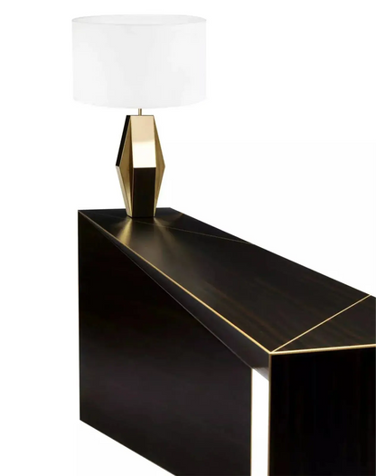 A sleek table lamp with a white shade and a geometric gold base sits on the Modu Console, which features a dark, angular design.