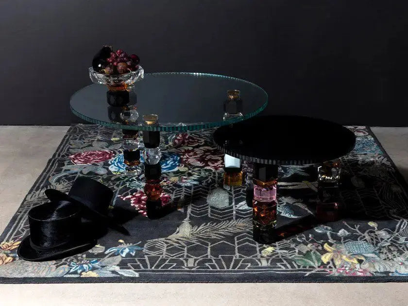 Two Orlando Crystal Coffee Tables with ornate legs rest on a floral-patterned rug, topped with a bowl of fruit. A hat and a cane are placed beside them.