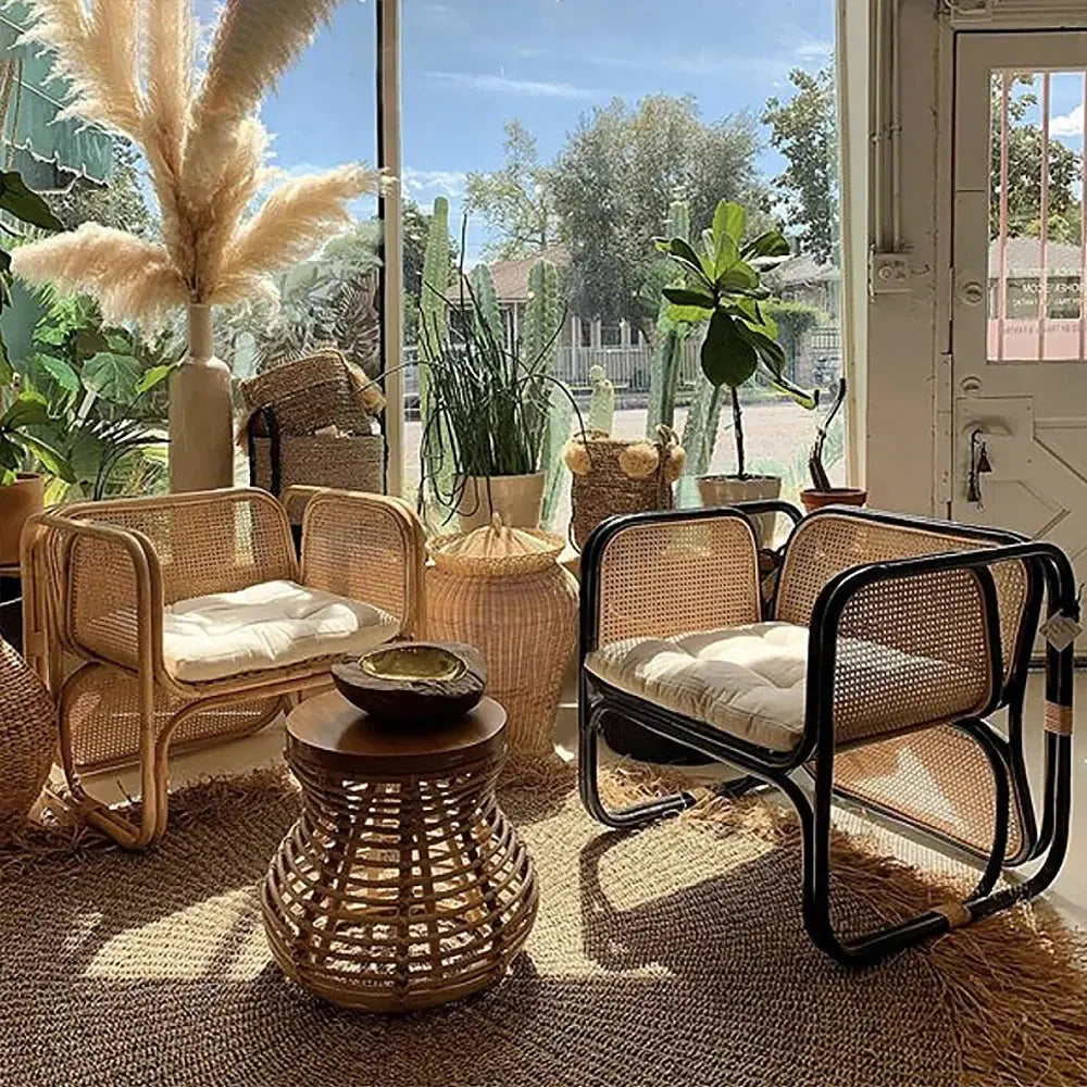 Two Morocco Style Rattan Armchairs with cushions are positioned around a small round wicker table in a sunlit room with large windows. The space is decorated with potted plants and pampas grass.