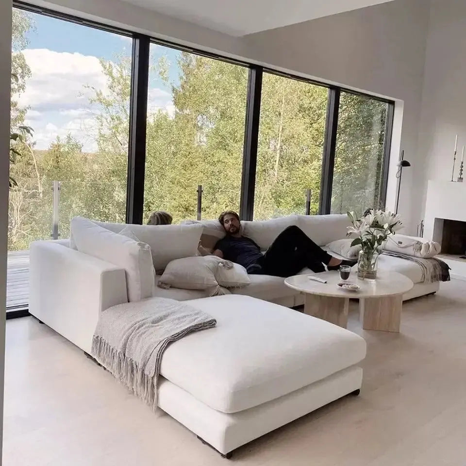 A man relaxes on The Heavenly White Modular Sectional in a bright living room with large floor-to-ceiling windows overlooking a lush green landscape. Nearby, a coffee table adorned with flowers and holding a glass of wine completes the serene setting.