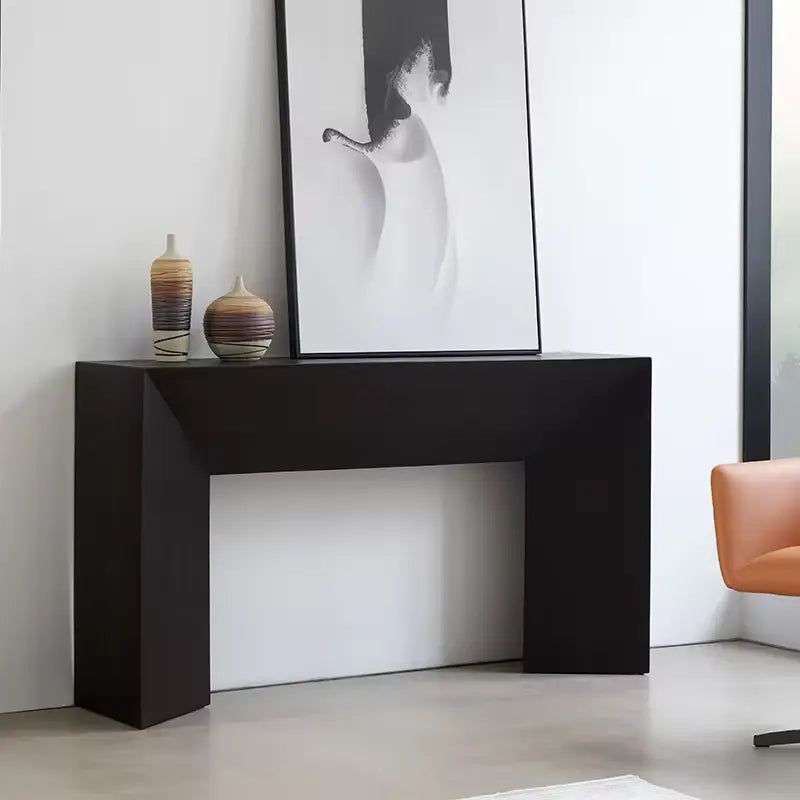 The Modu Console, a modern black table, stands against a white wall, showcasing a large abstract painting and two decorative vases. An orange chair is partially visible on the right.