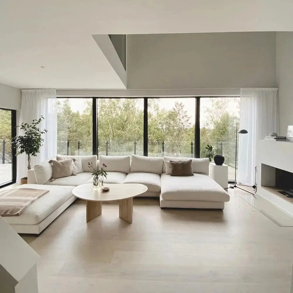 Modern living room with large windows featuring The Heavenly White Modular Sectional, a round wooden coffee table, a potted plant, and views of greenery outside. Neutral color scheme with minimalist decor.