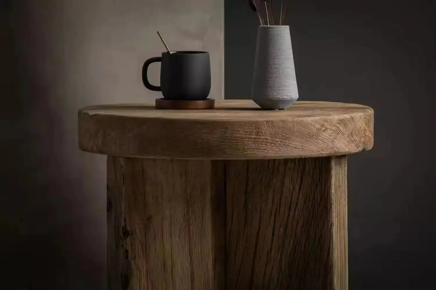 An Antique Round Wood Tea Table adorned with a black mug and a gray vase filled with reed sticks. The setting has a dimly lit, grayish background.