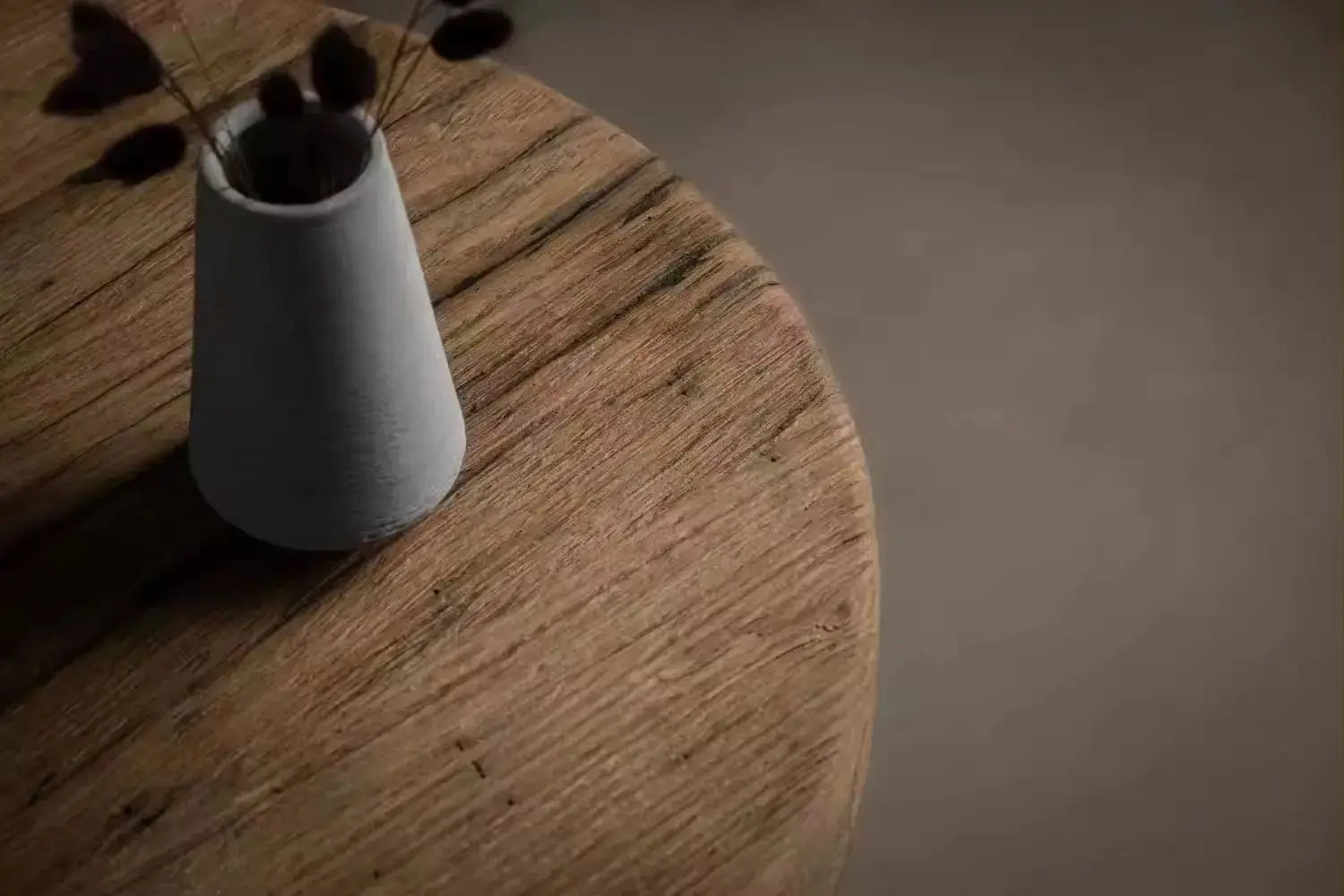 A minimalist white vase with dried flowers is placed on the Antique Round Wood Tea Table, showcasing its rich grain and texture against a plain background.