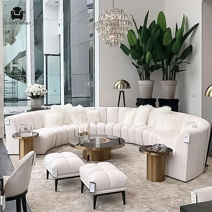 Luxurious living room featuring the Grand Curved Sectional sofa in white, round coffee tables, plush stools, tall greenery, a crystal chandelier, and floor-to-ceiling windows. Various price tags are visible on the furniture.