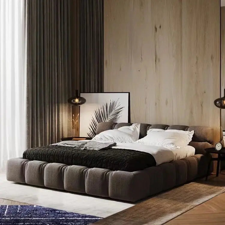 Modern bedroom featuring the Modern Zen Bed, a low, cushioned piece adorned with a black throw and white pillows. A framed leaf print leans against the wood-paneled wall, flanked by two lamps. Gray curtains frame a large window.