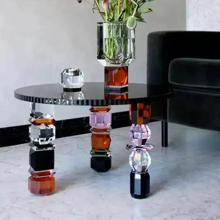 The Luxury Glass Coffee Table, featuring geometric, multicolored glass legs and a sleek black top, is adorned with a vase of flowers and a small decorative object. This elegant table is placed next to a dark armchair on a light floor.