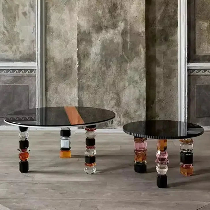 Two Luxury Glass Coffee Tables with black tops and legs composed of stacked, multicolored geometric shapes are placed on a floor against a textured wall with decorative moldings.