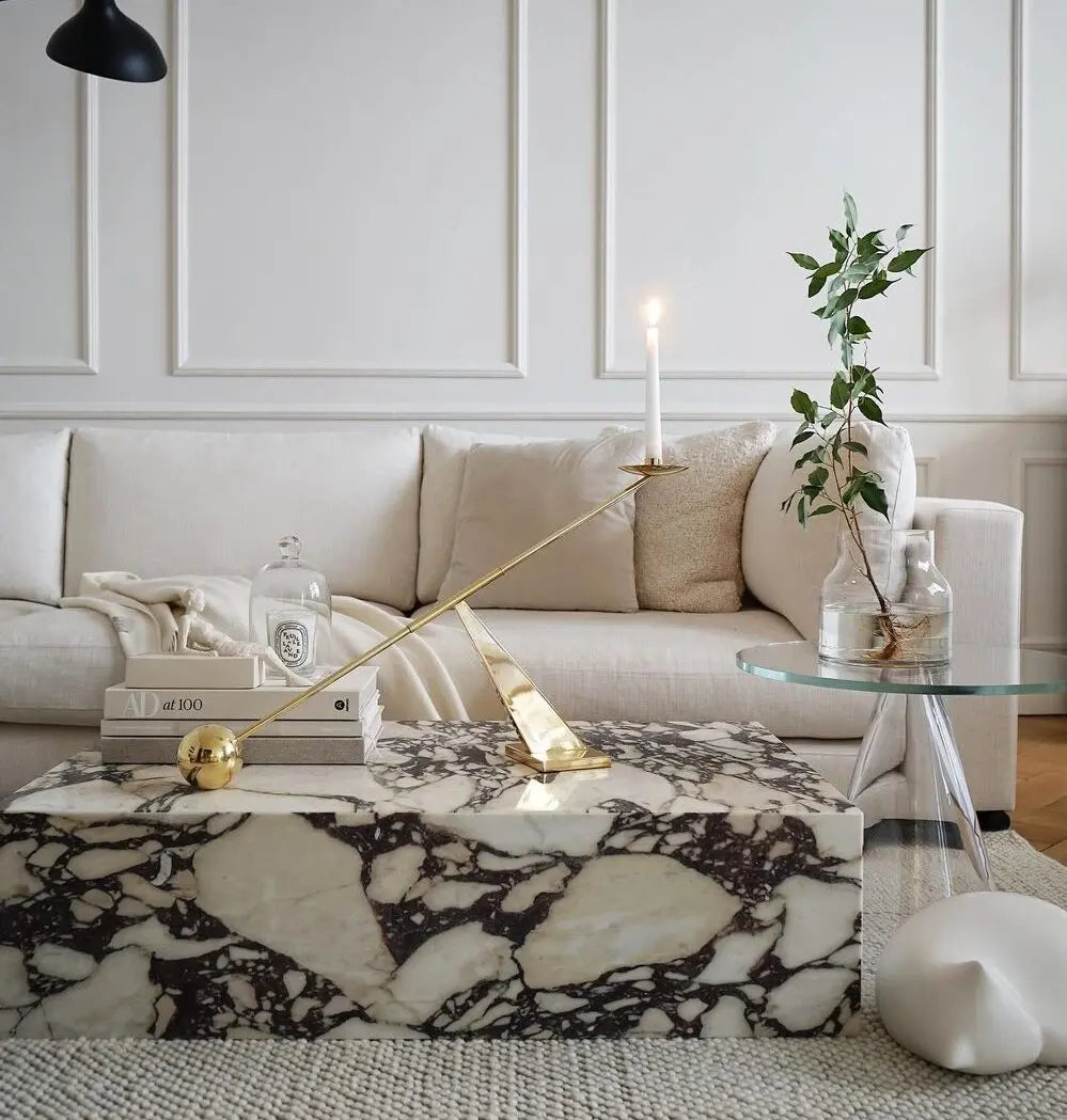 A modern living room featuring a beige sofa, the Calcutta Viola Marble Coffee Table, a decorative candle holder, and a stack of books. A glass side table holds a vase with green branches.