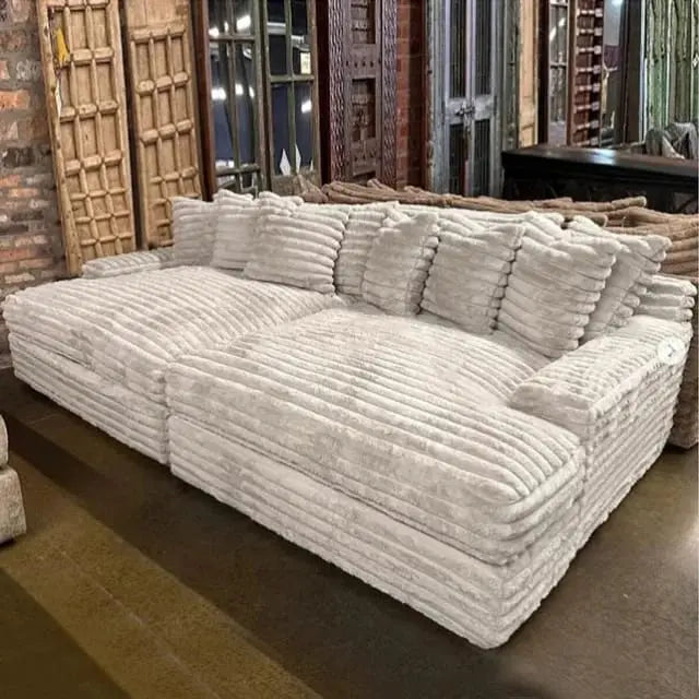 The Oasis Double Chaise, a large textured gray sectional sofa with multiple cushions, sits elegantly in a room adorned with various wooden decor and mirrored furniture pieces in the background.