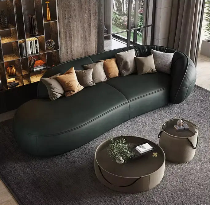 Modern living room featuring the Costera Sofa in dark green, elegantly curved and adorned with assorted throw pillows, complemented by round coffee tables, built-in shelving, and large windows providing a stunning outside view.