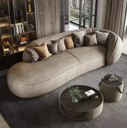 A modern living room featuring the Costera Sofa in a curved beige design, adorned with assorted cushions. The space includes two round coffee tables decorated with books and a plant, all set against a wall lined with shelves and large floor-to-ceiling windows.