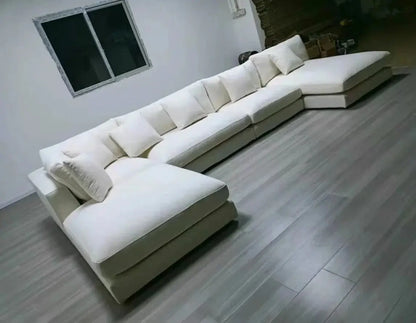 The Heavenly White Modular Sectional, a large off-white sofa with multiple cushions, is arranged in a spacious, minimally decorated room with light wood flooring and a window on the left wall.