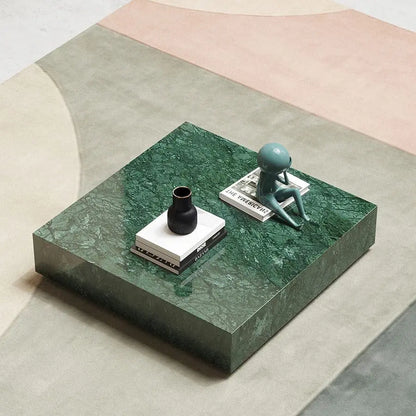 A Modern Green Marble Top Coffee Table featuring a black vase, two stacked books, and a small teal figurine sitting atop one of the books. The table is placed on a multicolored, geometric-patterned rug.