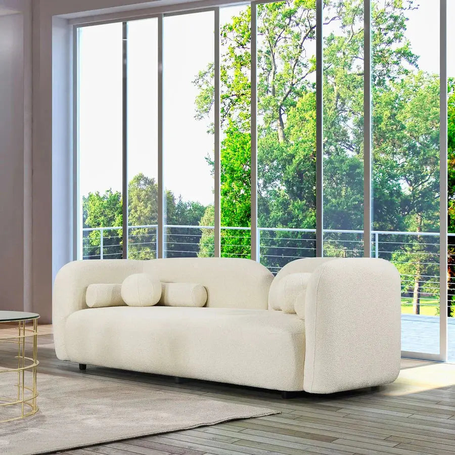 A modern Nordic White 3 Seater Sofa adorned with various pillows is placed near large windows that offer a view of a green, tree-filled landscape. A coffee table is partially visible on the left.