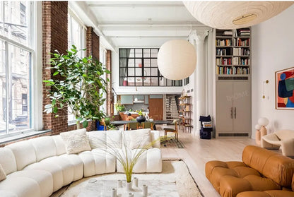 Bright, spacious living room featuring the Grand Curved Sectional, large windows, indoor plants, a marble table, bookshelf, and a hanging paper lantern. An open loft area and a dining space are visible in the background.