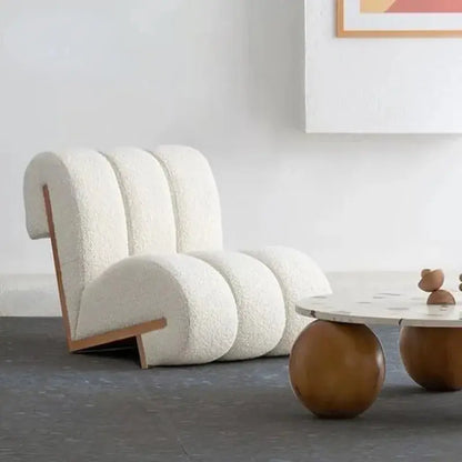 The Ella Leisure Chair, featuring a modern white plush design with unique curves, is situated near a round coffee table with wooden legs. A piece of abstract artwork is partially visible on the wall above.